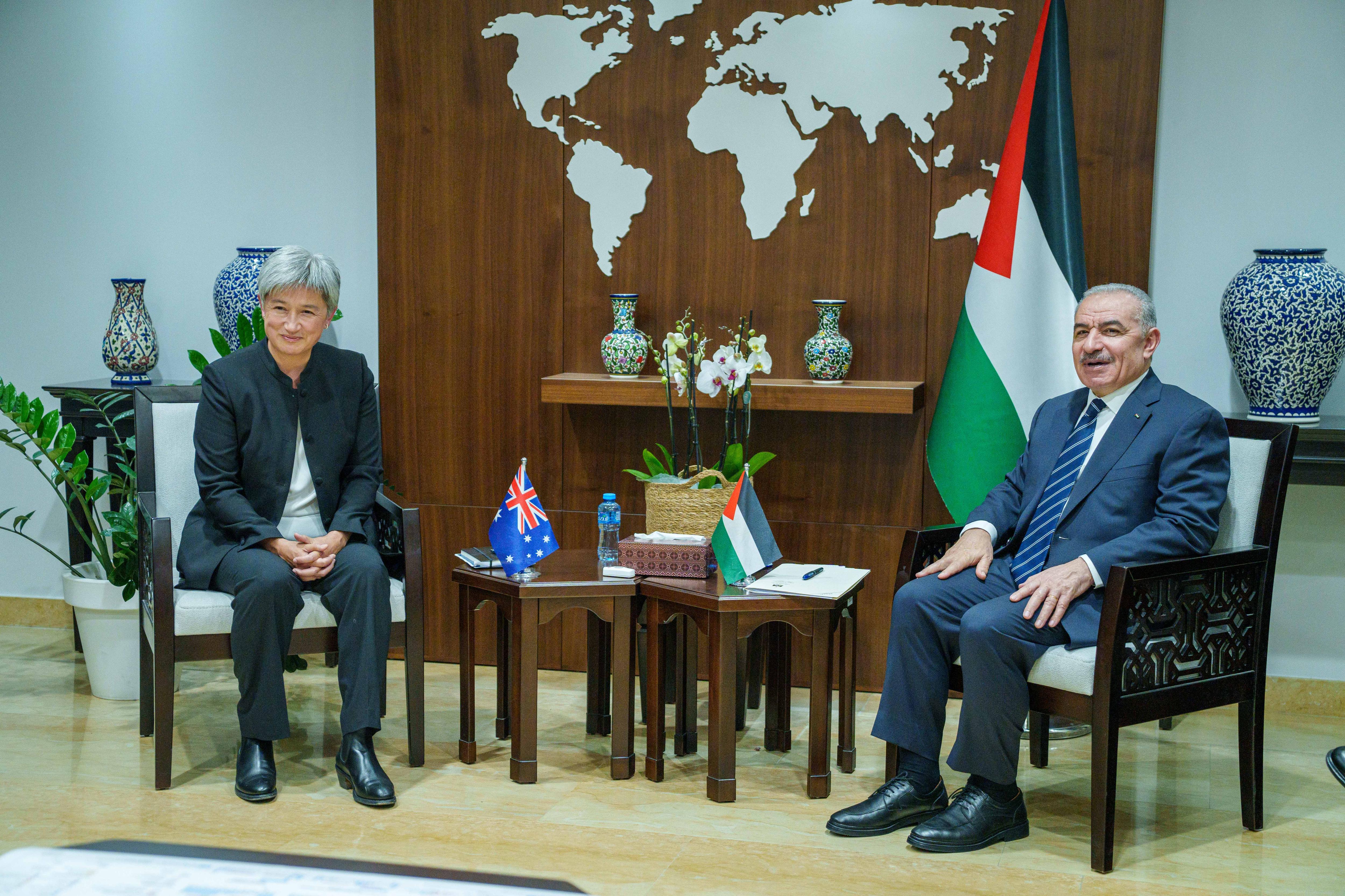 A medium shot of a woman and a man sitting in chairs in suits, next to their country's flag, smiling