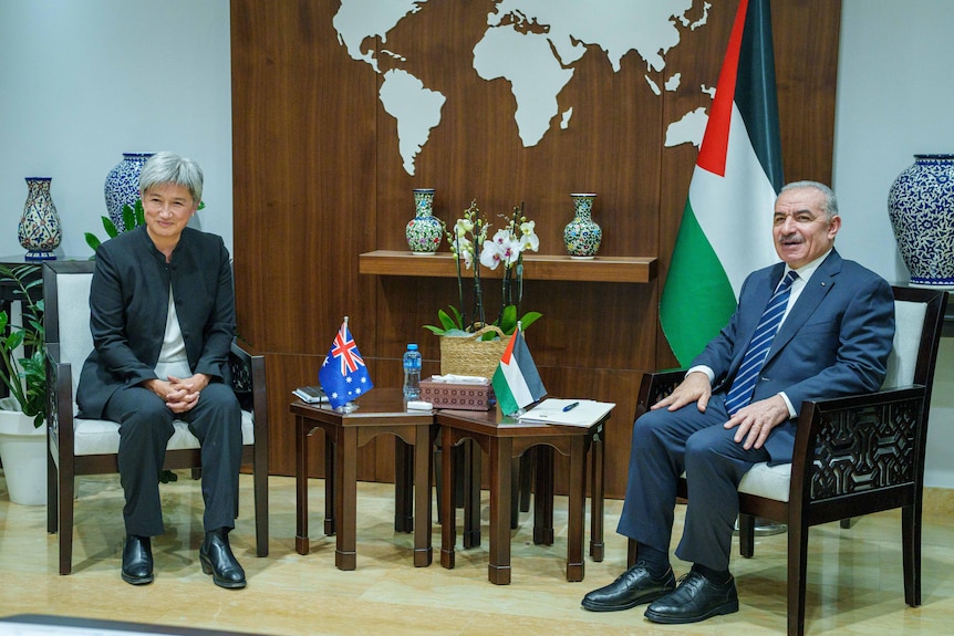 A medium shot of a woman and a man sitting in chairs in suits, next to their country's flag, smiling