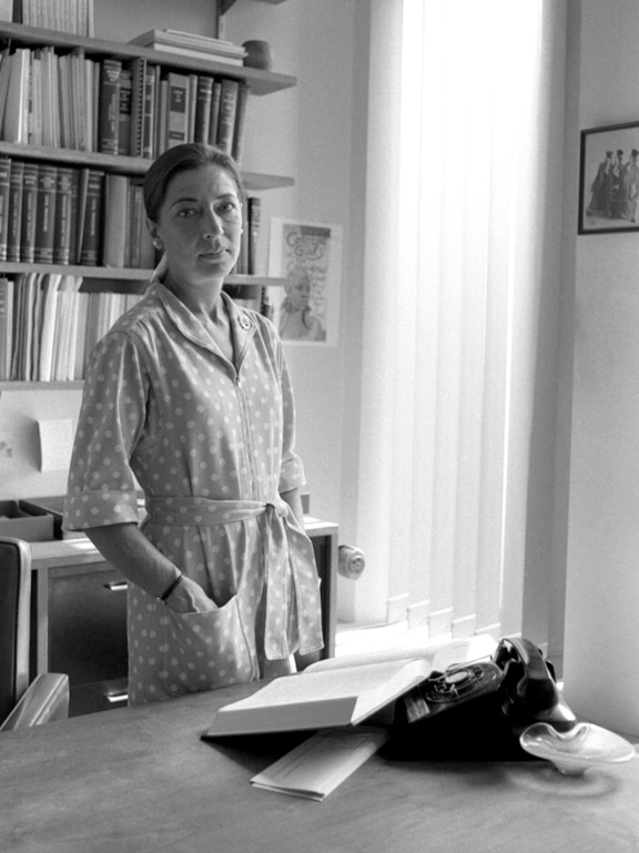 A black and white image of a young woman in an office with her hands in the pockets of her dress