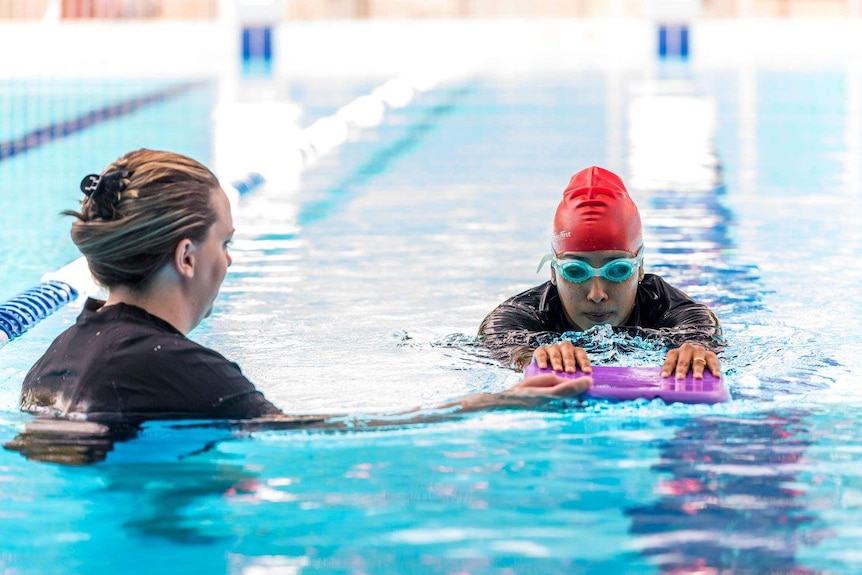 A woman with a kick board, swims in a pool with the assistance of a instructor.