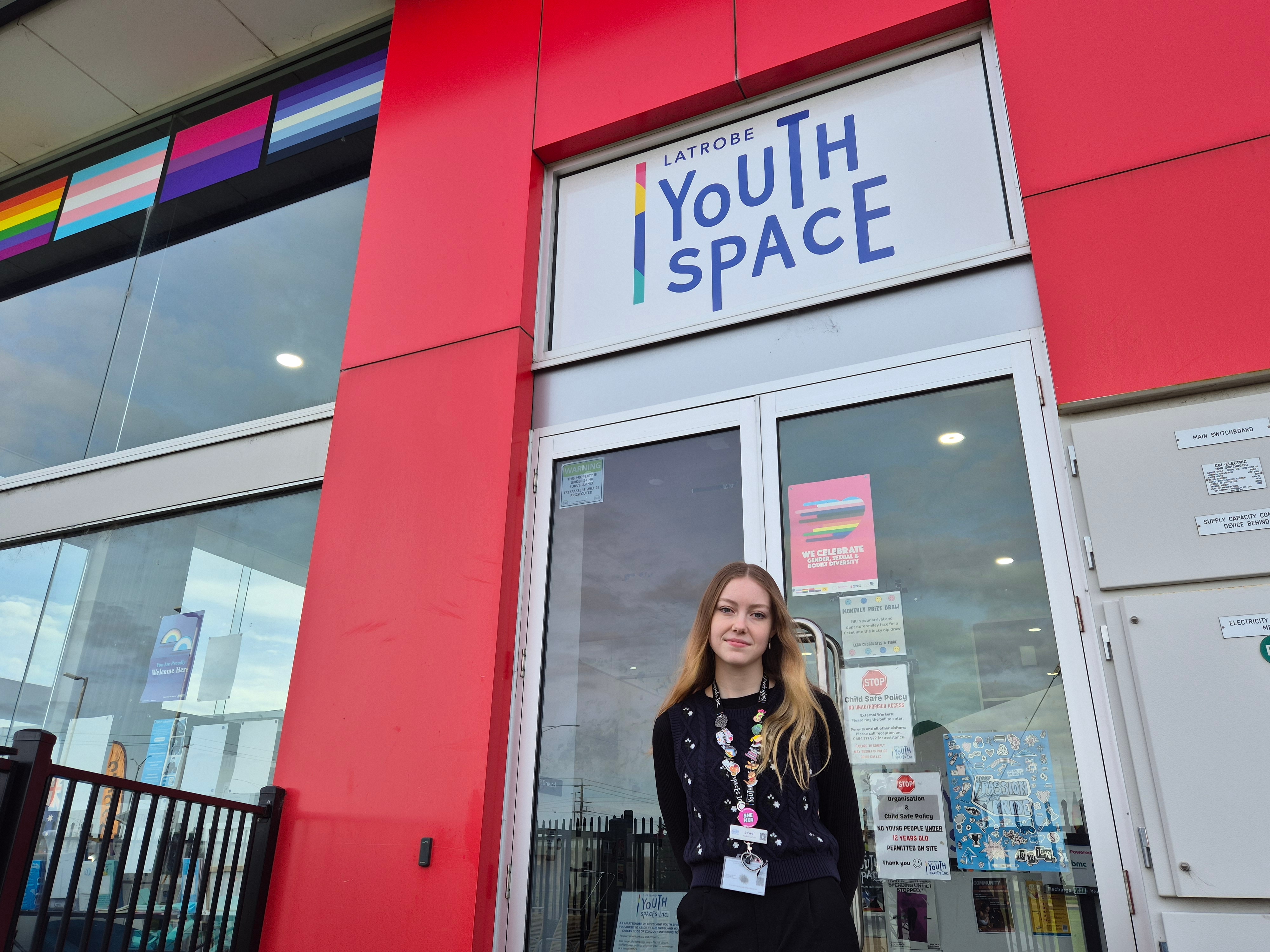 A young woman with long strawberry blonde hair in a dark top stands outside a colourful building with a Latrobe Youth Space sign