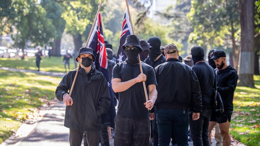 A group of men dressed in black with hats and masks hold Australian flags at an anti-immigration rally in Melbourne.