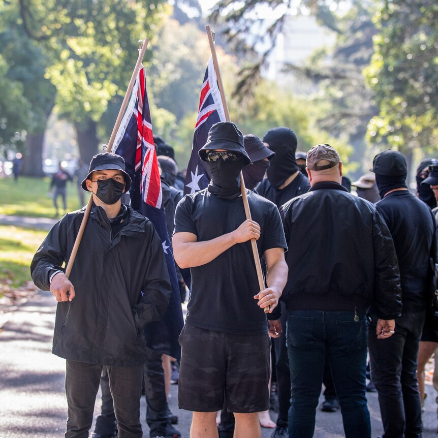 A group of men dressed in black with hats and masks hold Australian flags at an anti-immigration rally in Melbourne.