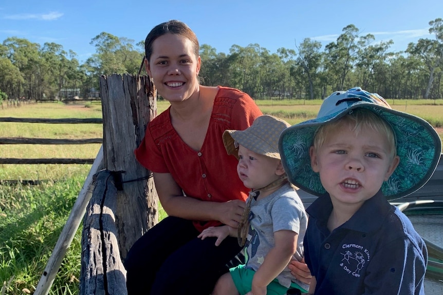 A woman with two young boys sit on the tray of a ute near a wooden fence in an open paddock.