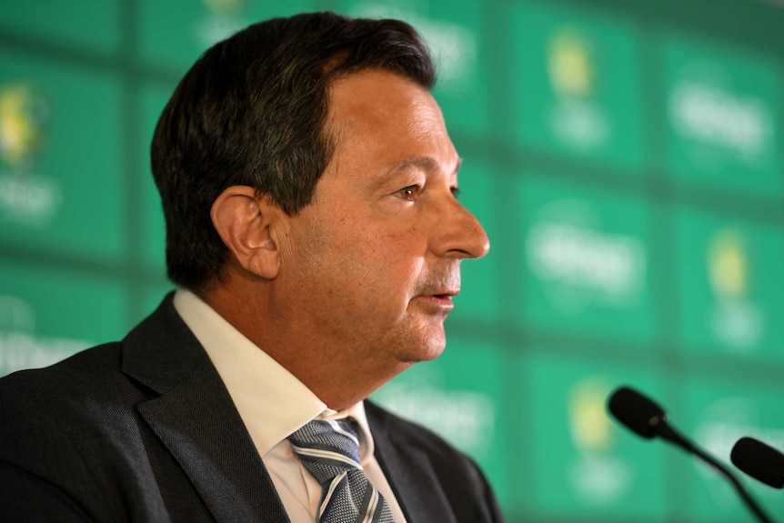 A side-profile view of a man sitting in front of two microphones wearing a suit and tie.