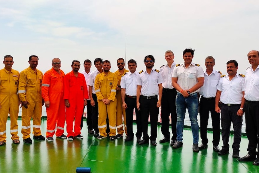 On an overcast day, you view a line of male seafarers pictured in a line onboard the green deck of a ship.