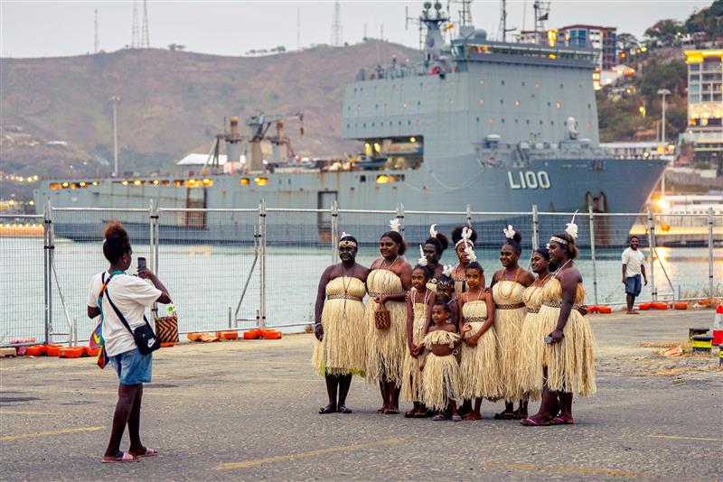 A woman takes a picture of a group of females dressed in traditional attire who are posing in front of a battleship