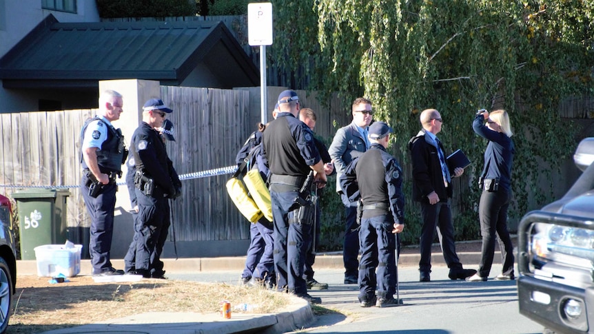 A group of 10 police officers stand on road near police tape, looking down the street or at each other.