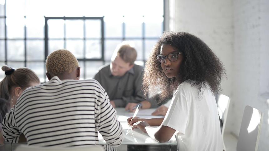 Woman looks at camera while colleagues work at table to depict talking about pay/salary to close gender and racial pay gaps.