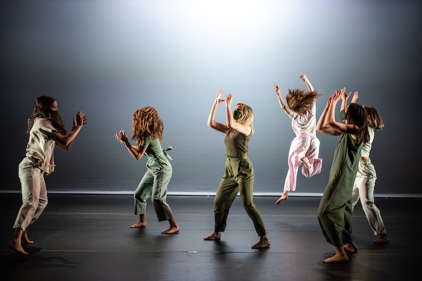 Six masked dancers wearing pastel green jumpsuits move energetically on a black stage with monochromatic light.