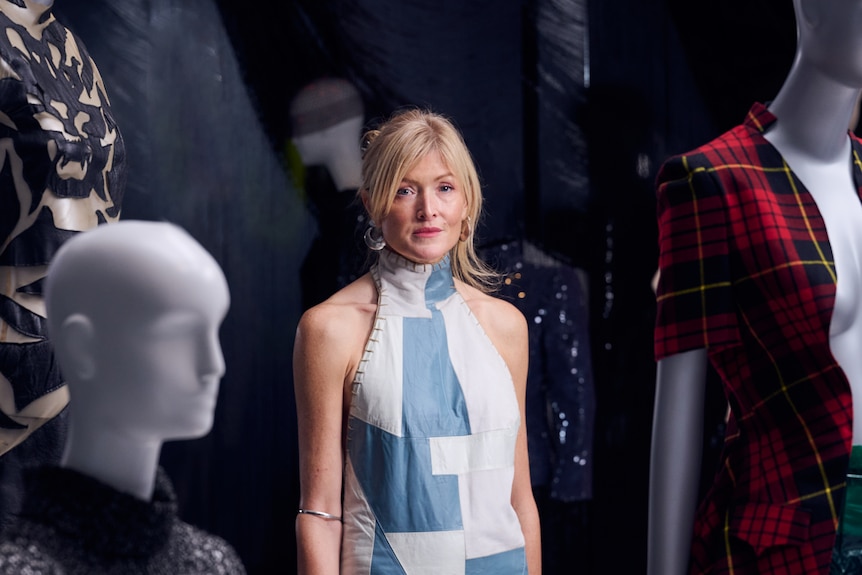 Middle-aged white blonde woman wears elegant blue and white halter dress among mannequins.