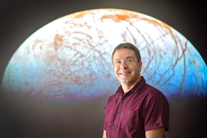 Astrophysics professor Jonti Horner smiles in front of a planetary backdrop