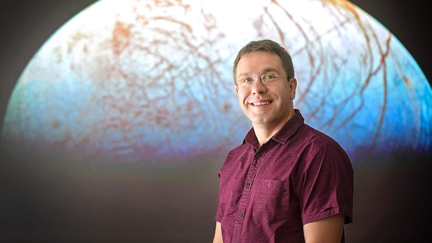 Astrophysics professor Jonti Horner smiles in front of a planetary backdrop