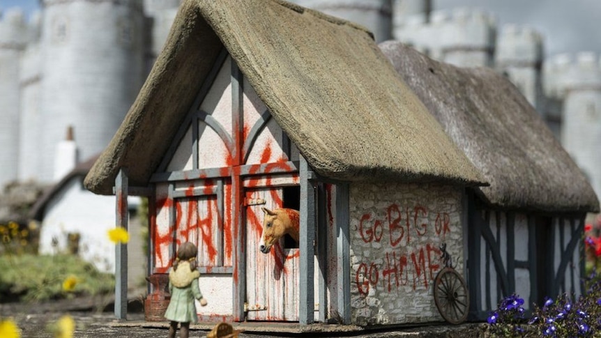 A figurine stands in front of a model building, which has been sprayed with the terms 'Banksy' and 'go big or go home'