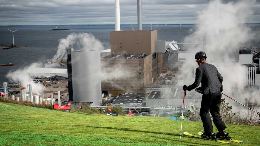 A person skis down what looks like a grass slope. In the background is a waste management centre.