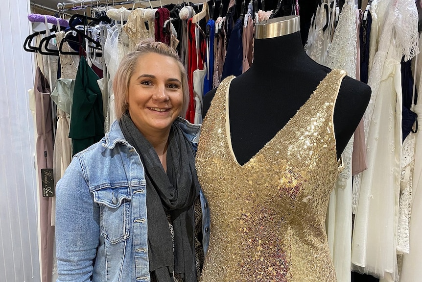 Young woman with short blonde hair smiling while standing amongst an array of formal dresses in her small Wagga Wagga studio.