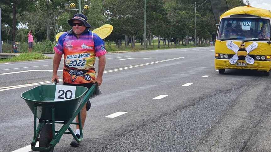 A man in a bee outfit pushing a wheelbarrow along the road
