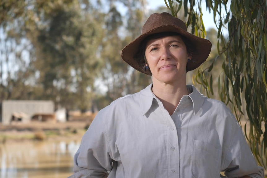 A woman is wearing a hat standing in front of a tree with a dam behind her.