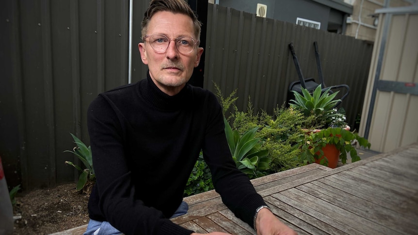 A man is wearing a black shirt and glasses and looks serious. There are plants in the background.