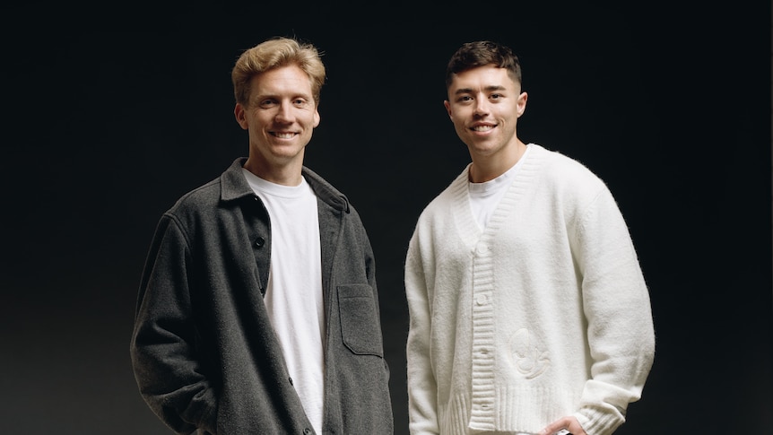 Brett wears a dark jacket and white shirt, Brad wears a white cardigan, in front of a black wall.