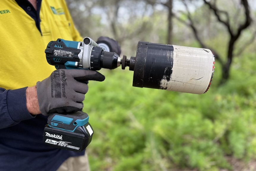 A gloved hand holds an electric drill with a hole saw attachment.
