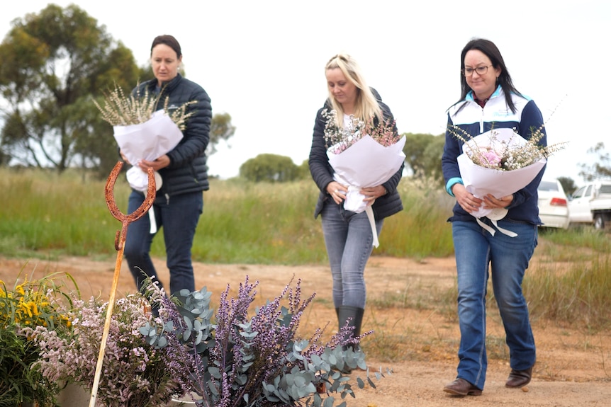 Three women holding bouquets of flowers walk to a place near a paddock to put the flowers.