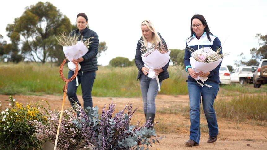 Three women holding bouquets of flowers walk to a place near a paddock to put the flowers.