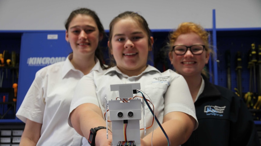 Three schoolgirls dressed in their uniforms look at camera, one of them holding aloft a small robot they've made.
