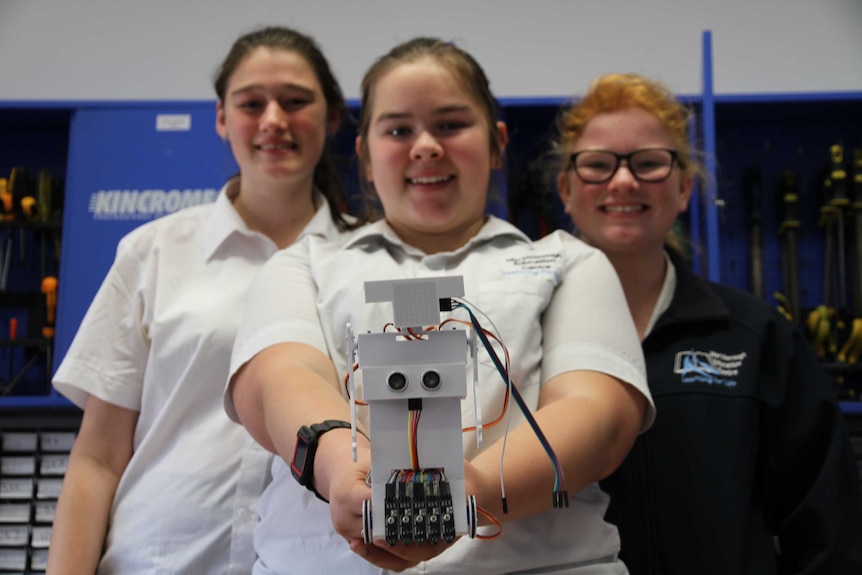 Three schoolgirls dressed in their uniforms look at camera, one of them holding aloft a small robot they've made.