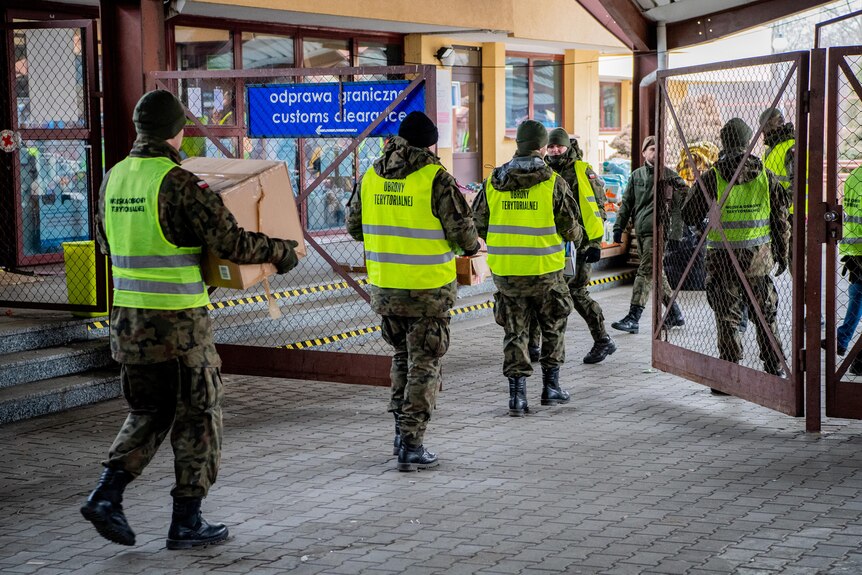 Men in army uniform with hi-vis vests over the top carry cardboard boxes through a gate