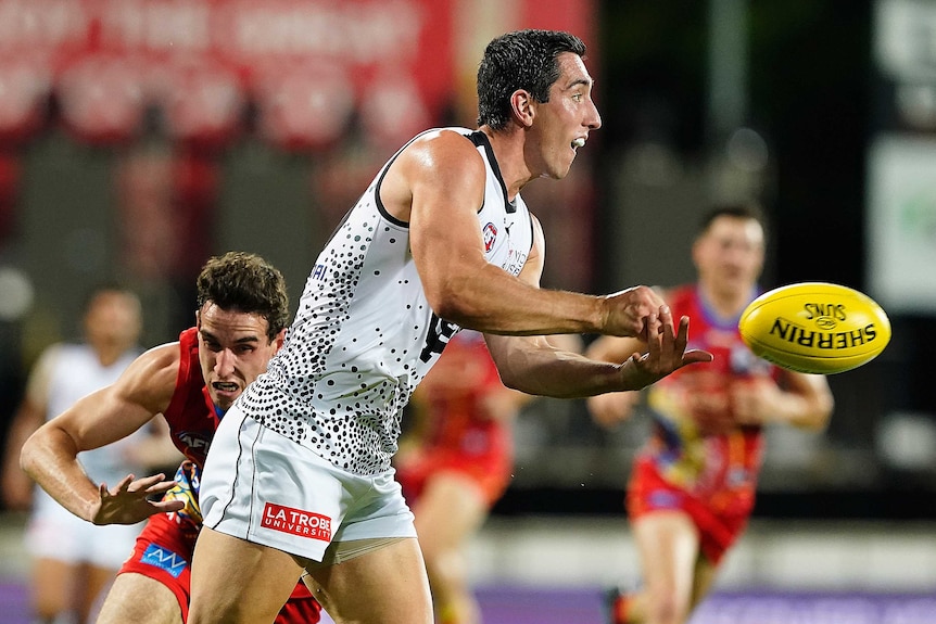 A Carlton AFL player handballs while a Gold Coast Suns opponent attempts to make a tackle.