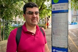 A man of Latin American background standing at a bus stop. He's wearing a red polo shirt and glasses