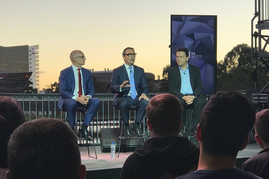 Three leaders on stage with Adelaide city buildings as a backdrop, nearing sunset.