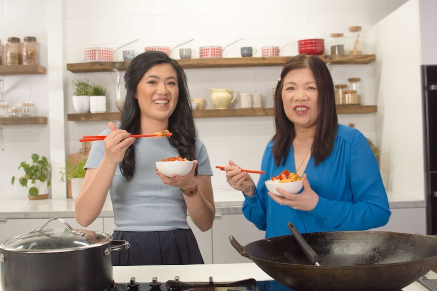 Annie Louey and her mum Jian Xian Louey stand in a kitchen, each holding a bowl of tomato and egg on rice.