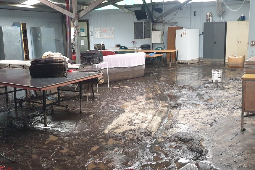 a common room in a workers' accommodation building is flooded with ankle-deep mud. Personal items are up on tabletops