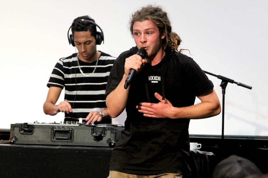 A man with dreadlocks talks into a microphone while another man in a striped T-shirt mixes on a desk behind him.