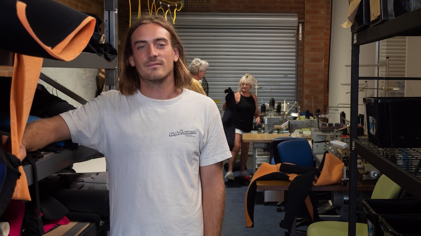 A man with long blonde hair and white t-shirt stands in a factory with two people behind him.