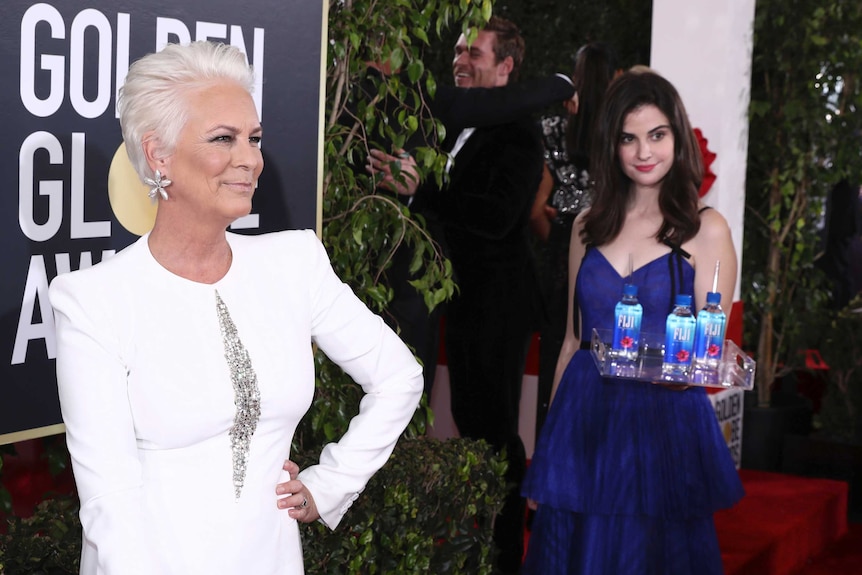 A woman holding a tray of water bottles stands in the background of a photo of Jamie Lee Curtis