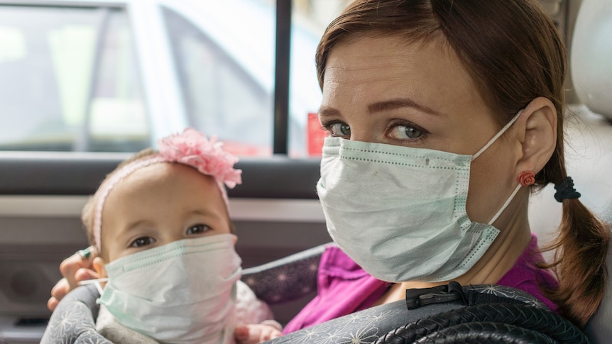 A white woman sitting in a car holding a young child, both wearing facemasks