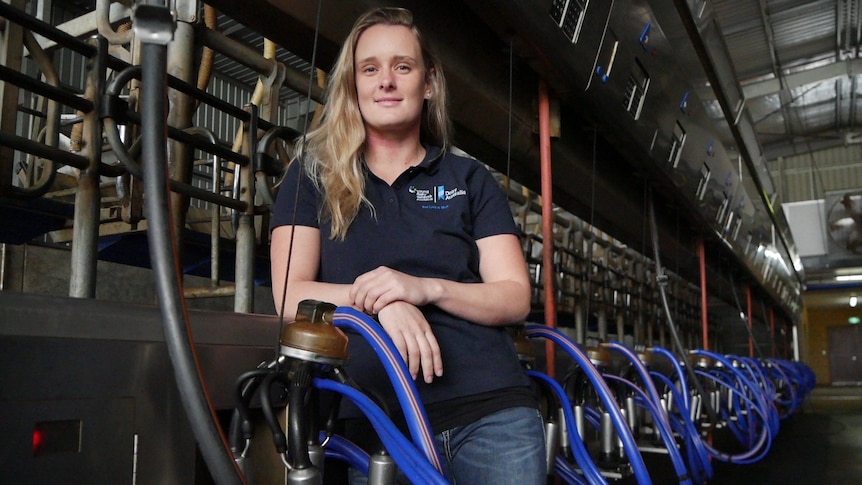 A tired-looking young, blonde woman in a navy polo shirt and jeans stands inside a dairy beside milking machines