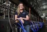 A tired-looking young, blonde woman in a navy polo shirt and jeans stands inside a dairy beside milking machines