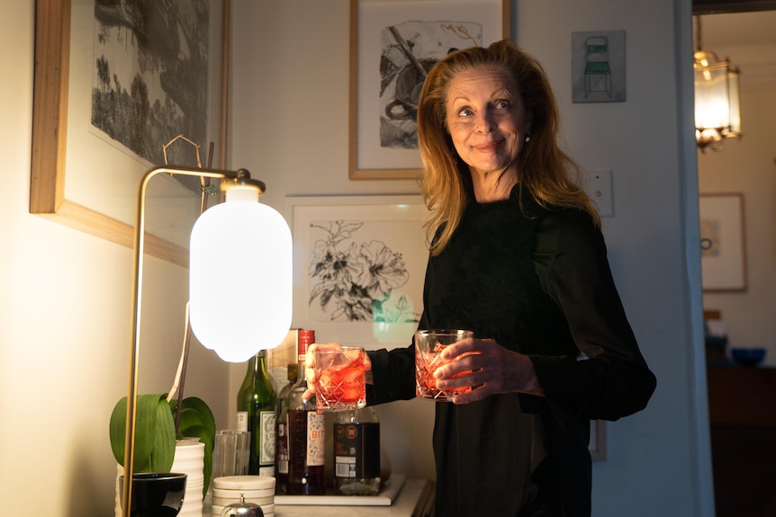 Heather Mitchell stands with two drinks in her hand in a room lit by a table lamp