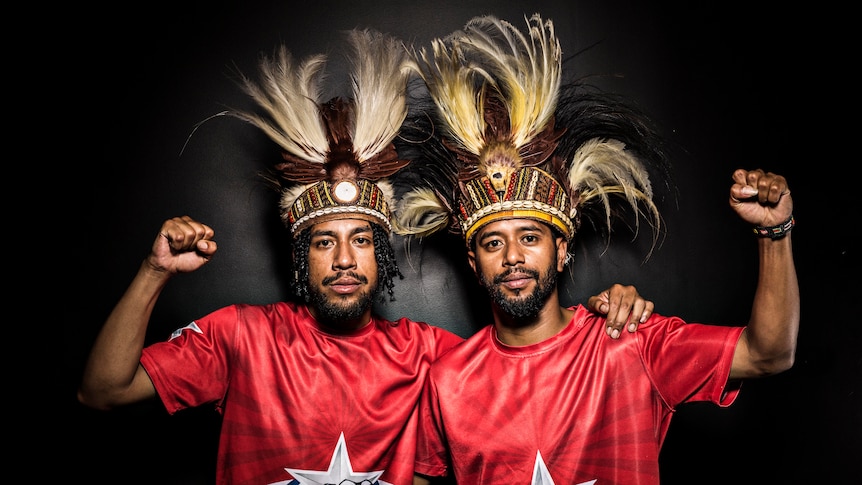 Brothers Yoshua and Sam Roem stand side by side against a black wall with fists raised in solidarity for West Papua.