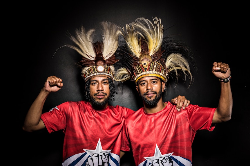 Brothers Yoshua and Sam Roem stand side by side against a black wall with fists raised in solidarity for West Papua.