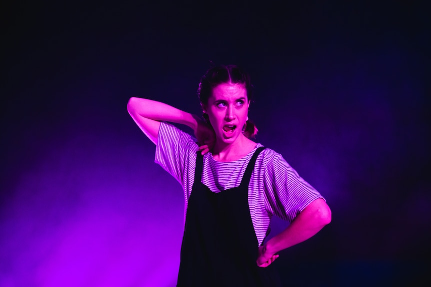 A brunette woman in her 30s, wearing overalls, bathed in purple light on stage, stands with her mouth open, one hand on her hip