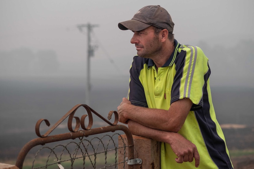 Craig McKimmie wears a high-vis yellow t-shirt, brown cap and leans against a gate, smoky background with burnt out paddocks.