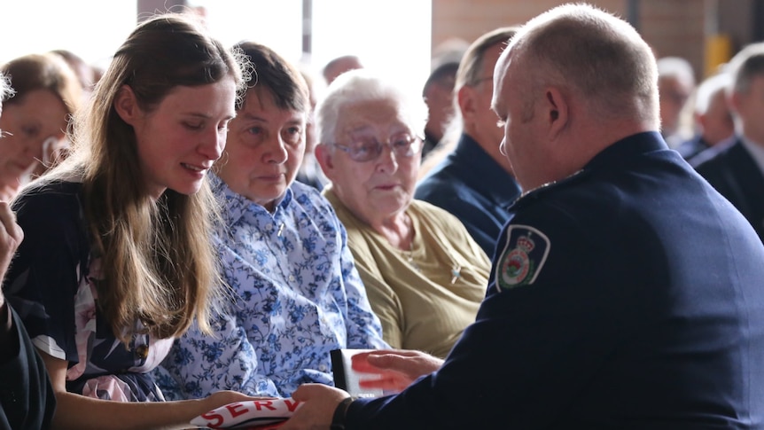 RFS chief Shane Fitzsimmons kneels in front of a seated and emotional Megan as he hands her her husband's awards.