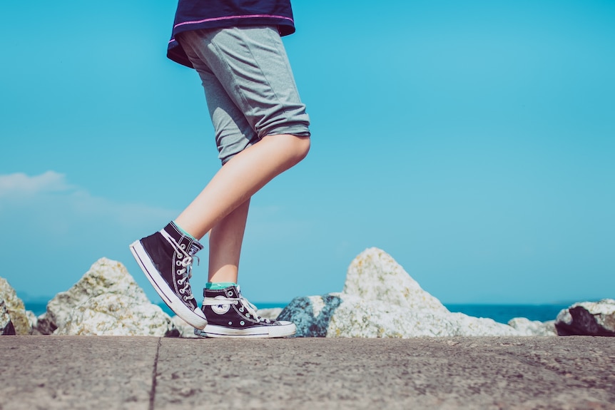 A child's legs and feet, wearing Converse hi-tops, walks along a concrete breakwall with the ocean in the distance