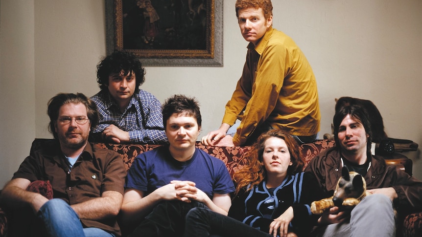 Six members of The New Pornographers sit on a couch and look at the camera