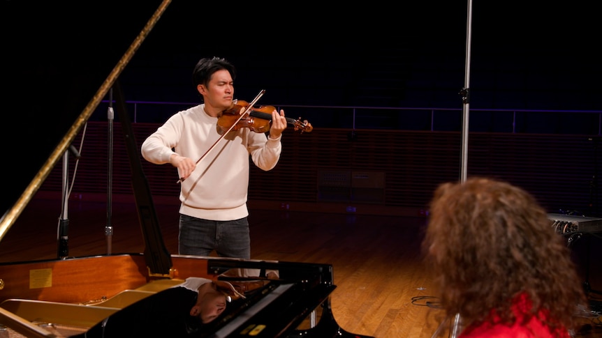 Tamara-Anna Cislowska from the side sitting at a grand piano. Ray Chen plays violin across from the bell of the piano.